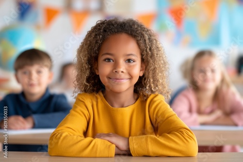 A smiling girl in a yellow sweater sits at a desk in a colorful classroom, with two other children blurred in the background.