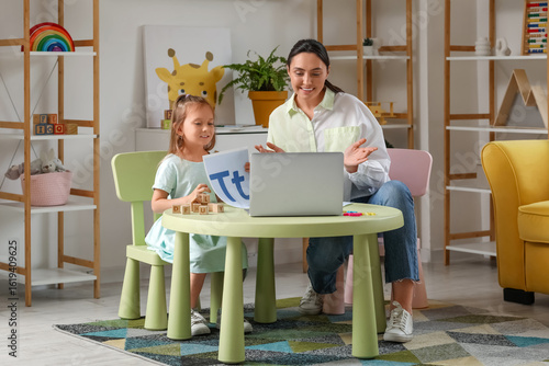Female speech therapist working with cute little girl and laptop at table in ...