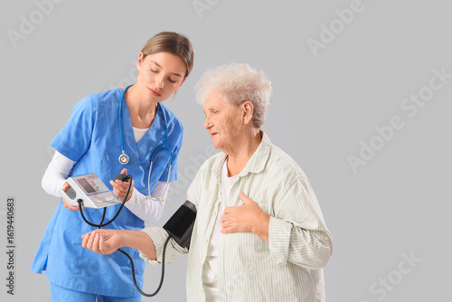 Senior woman with nurse measuring blood pressure on light background
