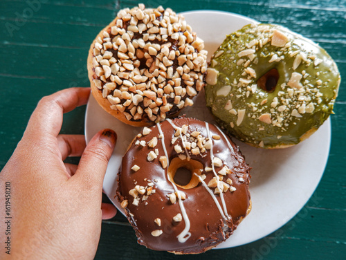 3 Donuts with varios flavour, chocolate, peanut and green tea donuts on the table, green background, top view, served by white plate, held by hand