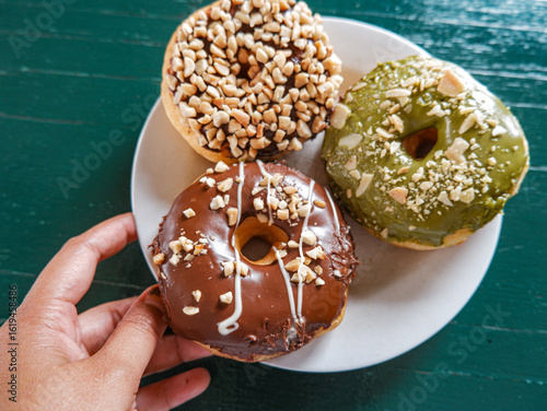 3 Donuts with varios flavour, chocolate, peanut and green tea donuts on the table, green background, top view, served by white plate, held by hand