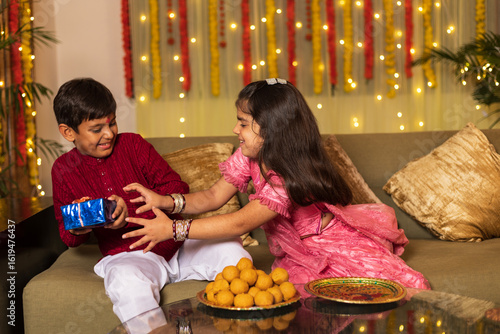 Indian brother and sister celebrating rakhi festival on the occasion of  Raksha Bandhan or bhaidooj with sweets and gifts while sitting on sofa and both wearing traditional festive cloths at home