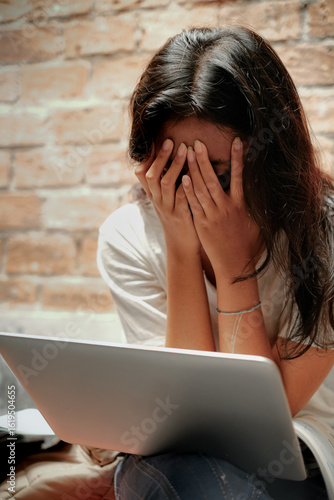 A young Asian teenage girl covering her face with both hands in front of a laptop while studying for high school final exam. Distress, frustration and mental health in education concept.
