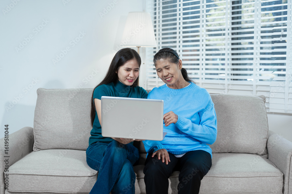 Fototapeta premium An Asian mother and daughter sit on a sofa, smiling as they look at a laptop screen. The warm indoor setting highlights shared learning, digital bonding, and meaningful family interaction at home