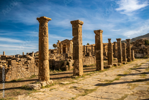 A striking low-angle shot showcases a series of remarkably preserved ancient Roman columns standing proudly against a dramatic blue sky with wispy clouds. These tall, weathered stone columns, with the
