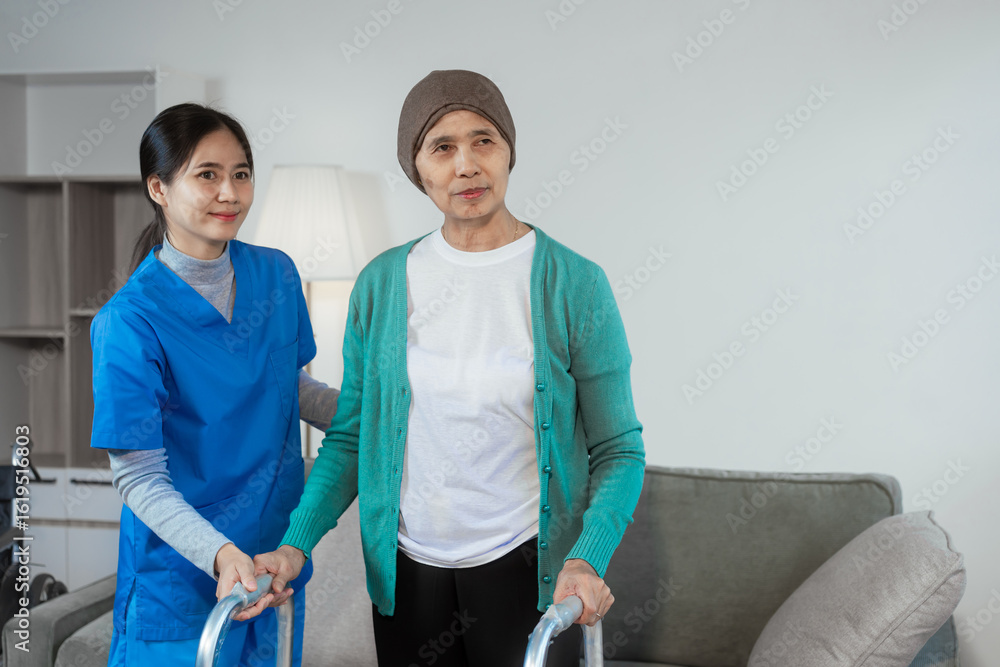 Fototapeta premium A young Asian nurse assists an elderly female cancer patient, wearing a headscarf, with using a walker. The patient sits on a sofa, preparing for rehabilitation and mobility training in a caring