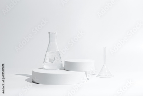 science with transparent glass erlenmeyer flask and funnel and beaker filled with colorless liquid water on a white cylinder base stand on desktop tabletop display with a simple clean background