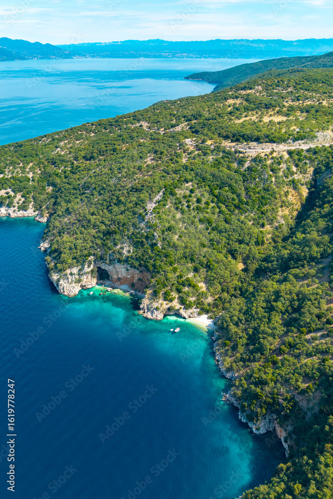 Fototapeta premium Aerial view of lush green hills and rocky coastline meeting turquoise waters on Cres Island. Secluded coves, boats, and a cave create a dreamy Mediterranean escape