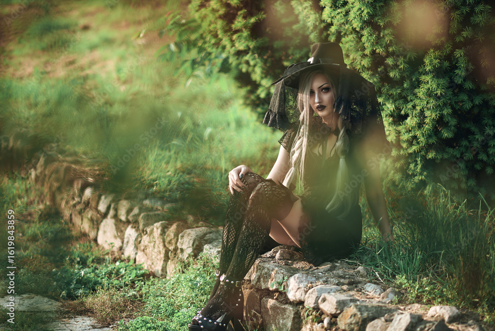 Naklejka premium Young woman in witch costume sits on stone path surrounded by greenery during daylight