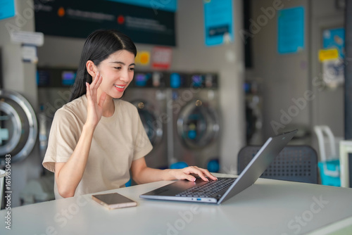 Confident woman focused on a virtual meeting using her laptop in a self-service laundry. Ideal for themes like flexible work and digital communication.