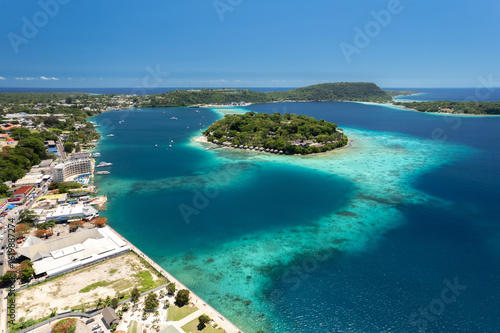 Aerial shot of Iririki Island in central Port Vila, Vanuatu. Island oasis set in a natural harbour with coastal cityscape and reef-lined shoreline visible.