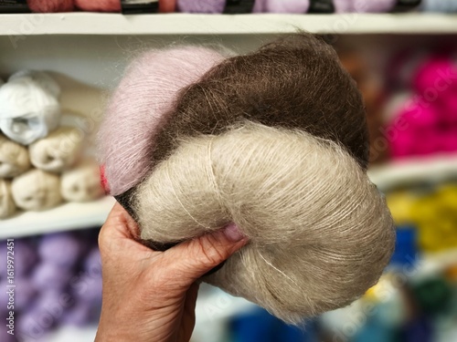 A hand holding soft mohair yarn in pastel pink, beige, and brown, with colorful yarn balls blurred in the background