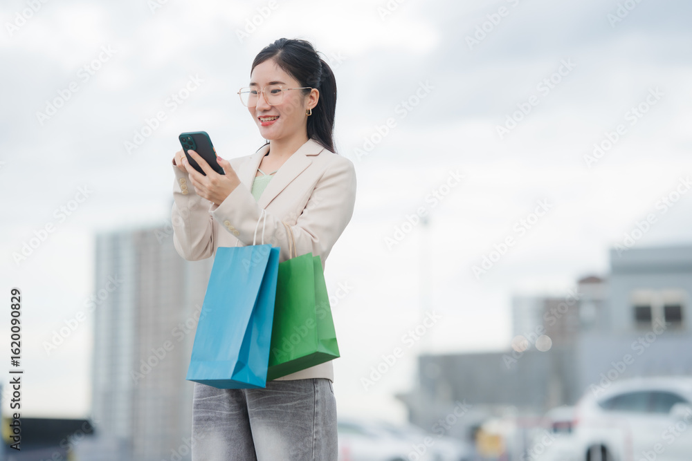 Fototapeta premium Shopping Delight: A stylish woman, clutching vibrant shopping bags, engages with her smartphone, showcasing the modern blend of shopping and technology in an outdoor setting.