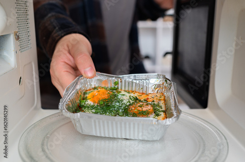 Hands place foil container with shakshuka lunch in microwave to heat.