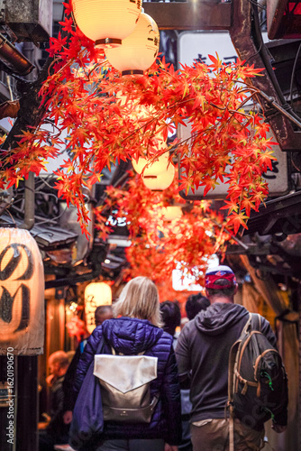 A narrow, lantern-lit alleyway in a Japanese town, filled with people and decorated with autumn leaves.