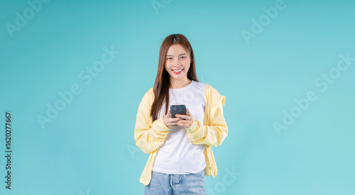 Happy young Asian woman using a smartphone while standing against a sky blue background, symbolizing modern communication, social media, digital lifestyle, and technology.