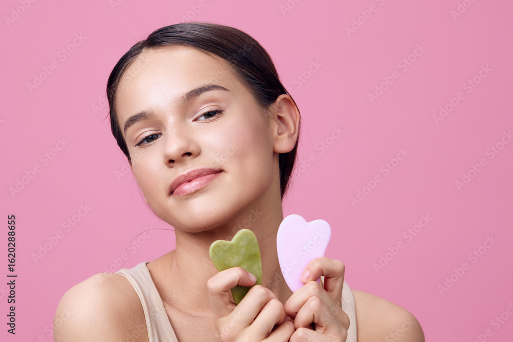 Young woman holding jade and rose quartz facial massagers, posing with a soft smile against a pink background, embodying a self care routine Young woman holding jade and rose quartz facial massagers, posing with a soft smile against a pink background, embodying a self care routine