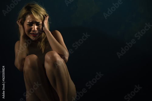 Distraught woman with blonde hair cowering in dark room while holding head in emotional pain and fear. Trauma and abuse concept