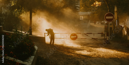Silhouetted worker in foggy construction site at sunrise with orange traffic sign