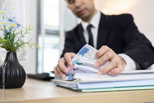 A man in a suit is working in an office. He is checking information and searching through a pile of documents, talking on his mobile phone at a table with a computer and keyboard in front of him.