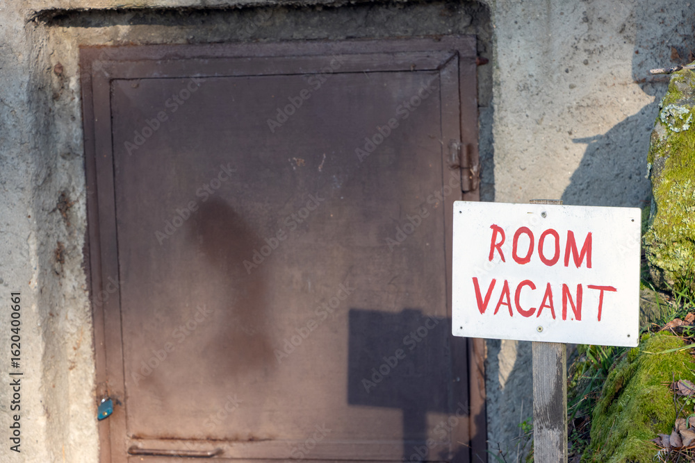 A white sign displaying 'Room Vacant' placed near a locked metal door A white sign displaying 'Room Vacant' placed near a locked metal door