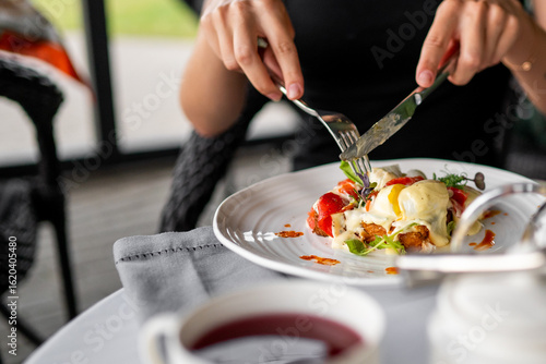 Photos Hands cutting eggs benedict on a plate with hollandaise sauce, garnished with greens, served with tea on a casual dining table—fresh and appetizing breakfast scene
