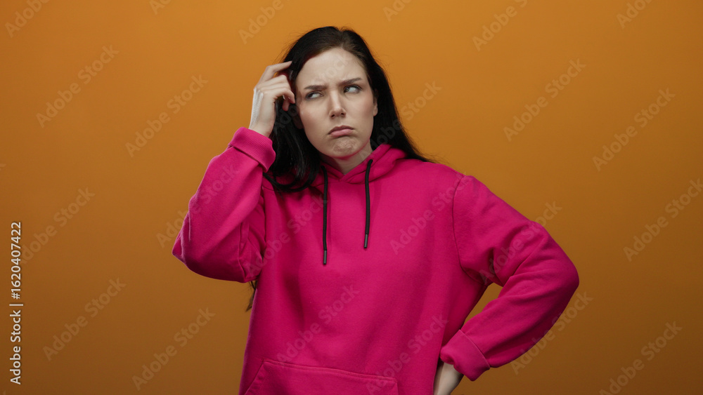 Fototapeta premium Woman contemplating while wearing a pink hoodie against an orange background, expressing deep thought with a pensive facial expression.