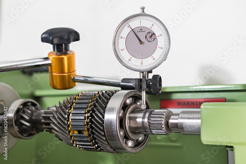 Gearbox shaft run-out measuring by dial indicator in holder on white background. Closeup a test probe and transmission gear mechanism with ball bearing and synchronizer rings clamped in lathe machine.