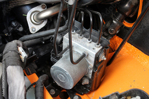 Car ABC system. Close-up of a vehicle ABS pump module with black hydraulic lines, silver aluminum block, and orange-painted engine bay background, highlighting automotive braking components