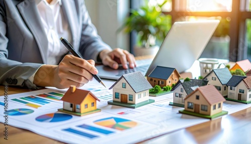 A businessperson analyzes real estate data with miniature houses, charts, and a laptop on a desk in a bright office.