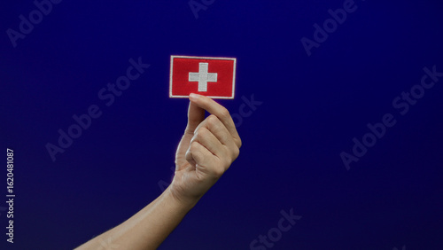 Fotografi Man holding a swiss flag embroidery patch against a blue isolated background, capturing a symbolic gesture with focus on nationality and culture