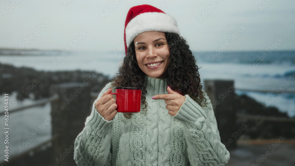 Obraz premium Woman wearing santa hat points at red mug by the beach, showing joy by the sea in a casual sweater, blending holiday cheer with coastal relaxation.