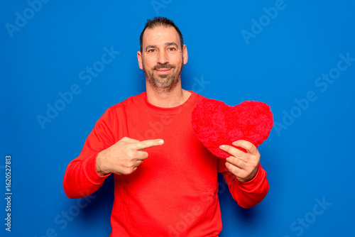 Bearded Hispanic man in his 40s wearing a red sweater holding a red heart as a symbol of romance and health, looking surprised and pointing at himself with his finger. Isolated on a blue background.