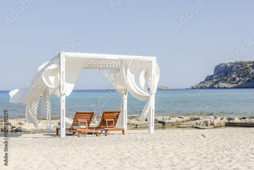 View of sun-drenched beach cabana with billowing white curtains frames a serene turquoise sea, inviting relaxation and escape, Lindos, Rhodes, Greece.