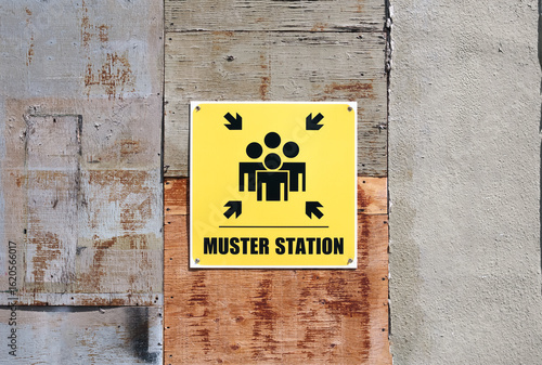 Yellow muster station sign on boarded up wall. Large sign with arrows and group of people. Assembly area for disaster response, fire drill or construction sites. Selective focus.