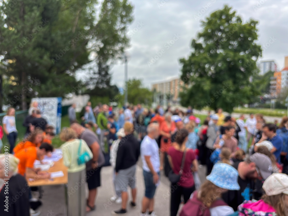 Out-of-focus photo showing a large, blurred crowd gathered at an outdoor event near a registration table in an urban setting. Out-of-focus photo showing a large, blurred crowd gathered at an outdoor event near a registration table in an urban setting.