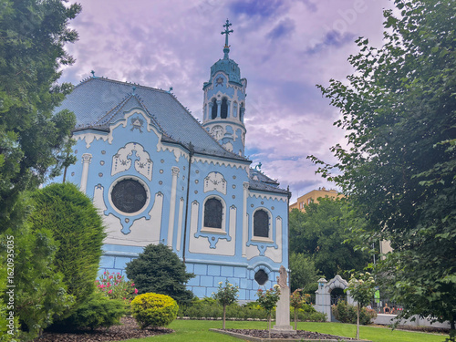 Photography Scenic view of the iconic Blue Church in Bratislava, Slovakia, surrounded by greenery and dramatic purple-tinted clouds