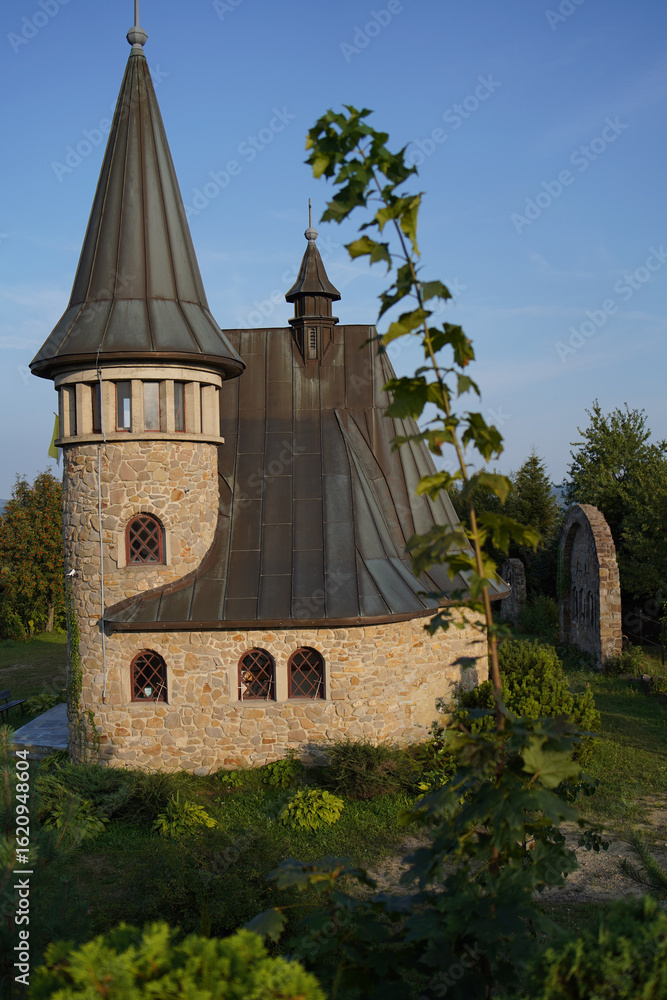 Fototapeta premium a stone church with a tall steeple and a clock on it. The church has a sloped roof and is situated on a grassy hill.