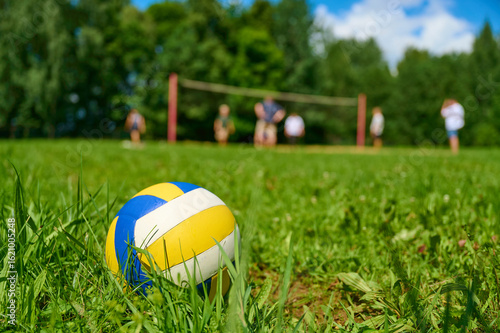 Close-up of a volleyball lying on lush green grass with a volleyball court and players in the background, under a blue sky with scattered clouds, capturing a lively outdoor sports scene