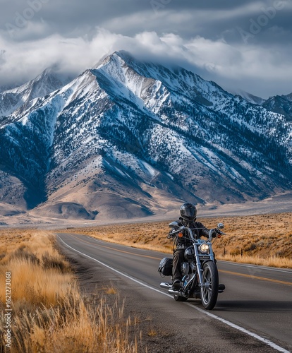 A motorcyclist rides down a scenic road with snow-capped mountains in the distance. The open road and dramatic landscape evoke a sense of freedom and adventure.