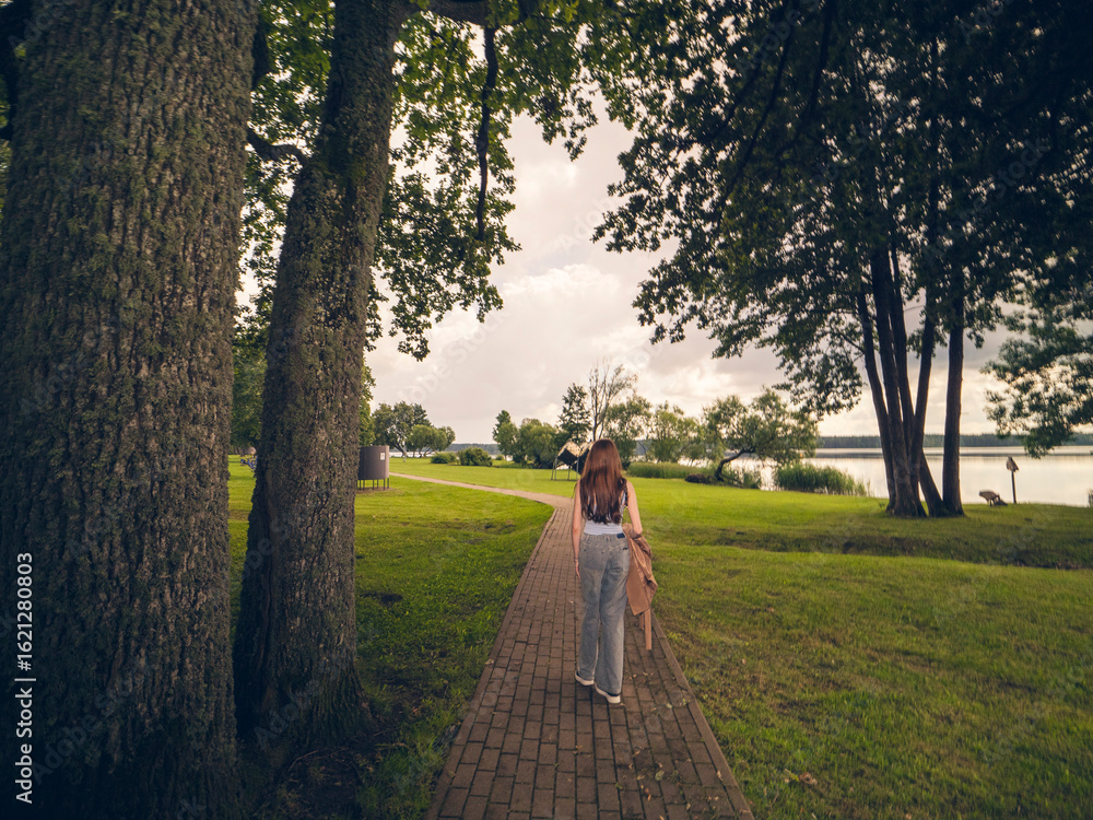 Fototapeta premium A woman walks down a path in a park. The path is lined with trees and the sky is cloudy. there is lake on the right and cloudy sky. Going on a blind date in a public place concept.