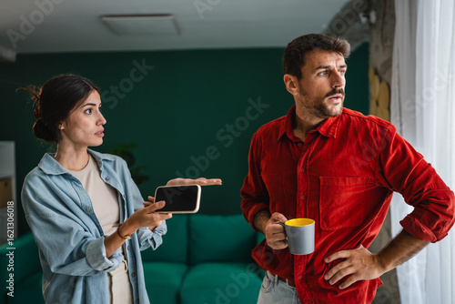 Photos Young couple arguing at home holding smart phone and coffee cup