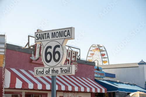 Iconic Route 66 ‘End of the Trail’ sign at Santa Monica Pier with Ferris wheel and striped canopies in the background