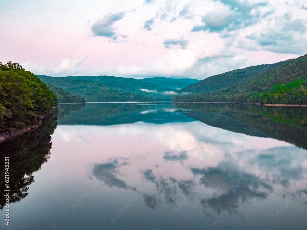 Fototapeta premium Dramatic sky over calm lake, with reflections of forest covered hills and clouds in the water, purple- late afternoon sky and mist in the distance. Pepacton Reservoir, Catskills, New York State USA.