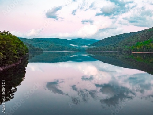 Dramatic sky over calm lake, with reflections of forest covered hills and clouds in the water, purple- late afternoon sky and mist in the distance. Pepacton Reservoir, Catskills, New York State USA.
