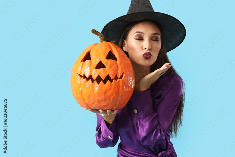 Fototapeta premium Beautiful young happy Asian woman dressed as witch for Halloween with carved pumpkin blowing kiss on blue background