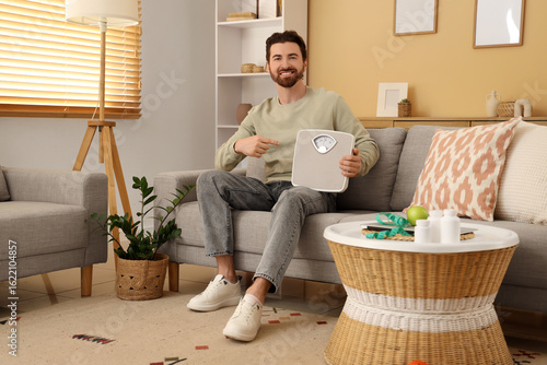 Young man with weight scales sitting on sofa at home