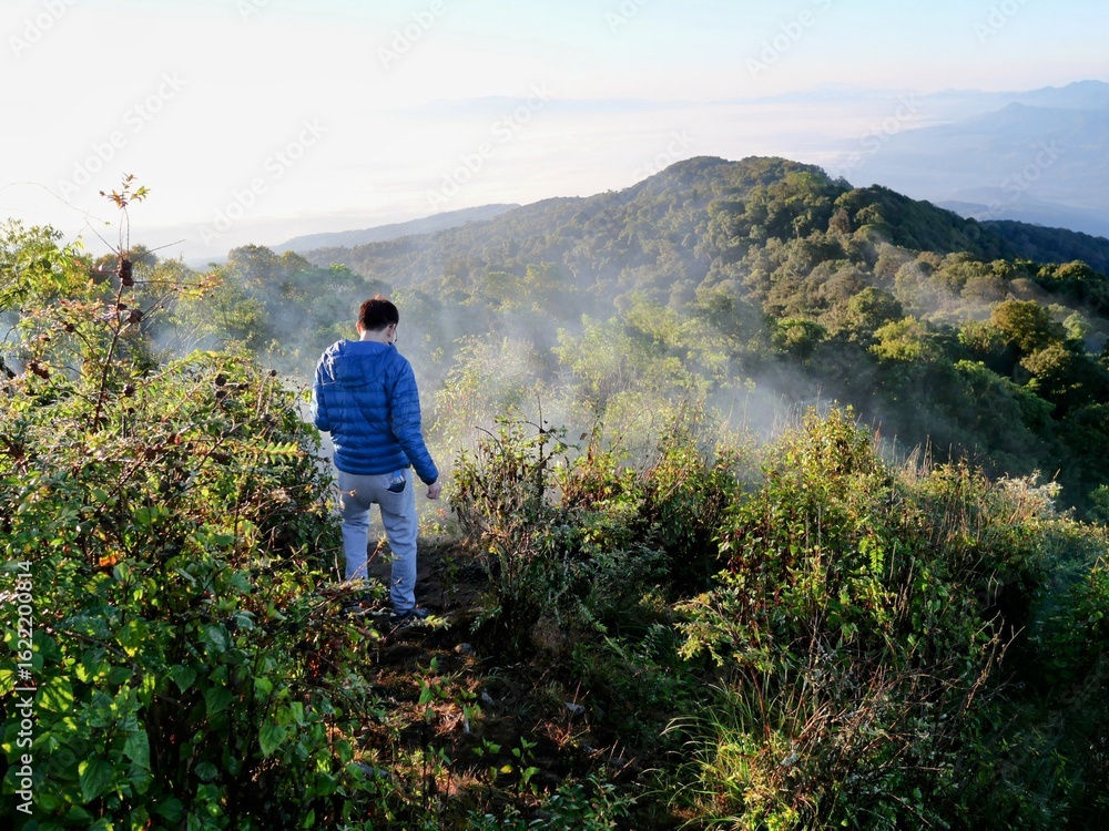 Naklejka premium Person in blue jacket overlooking misty hills at Doi Pha Hom Pok National Park, Thailand. Serene mountain landscape with lush greenery and tranquil atmosphere.