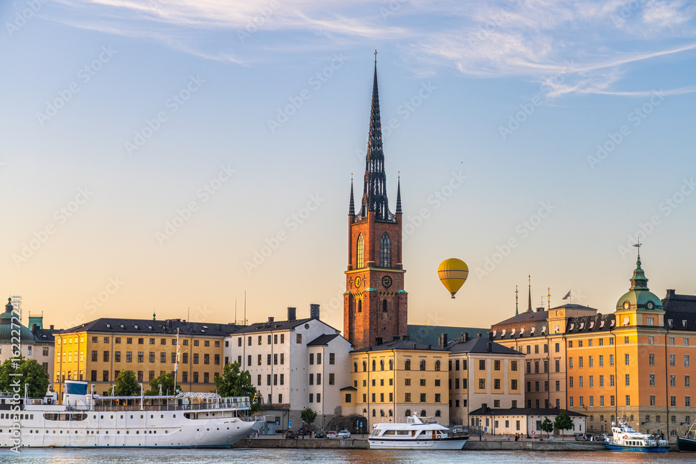 Fototapeta premium View of the Riddarholmen Church's spire piercing the skyline amidst historical buildings and a floating hot air balloon, Stockholm, Sweden.