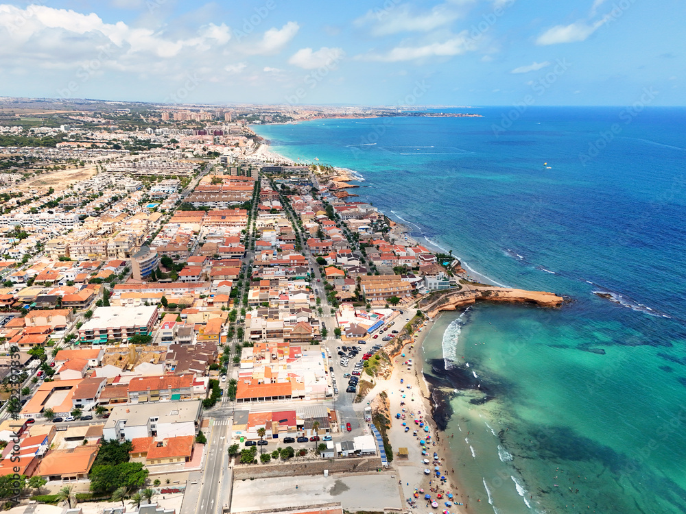Naklejka premium Aerial panorama of Torre de la Horadada on the Costa Blanca, showing colorful rooftops, sandy beach, turquoise Mediterranean waters, rocky coastline, and vibrant summer atmosphere